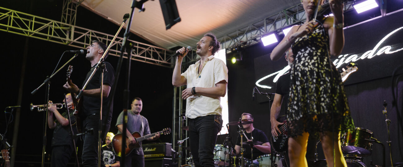 Musicians perform on stage during a lively summer festival at night, featuring vibrant lights and an enthusiastic audience enjoying the entertainment.