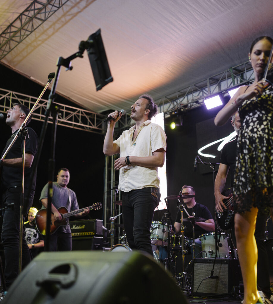 Musicians perform on stage during a lively summer festival at night, featuring vibrant lights and an enthusiastic audience enjoying the entertainment.